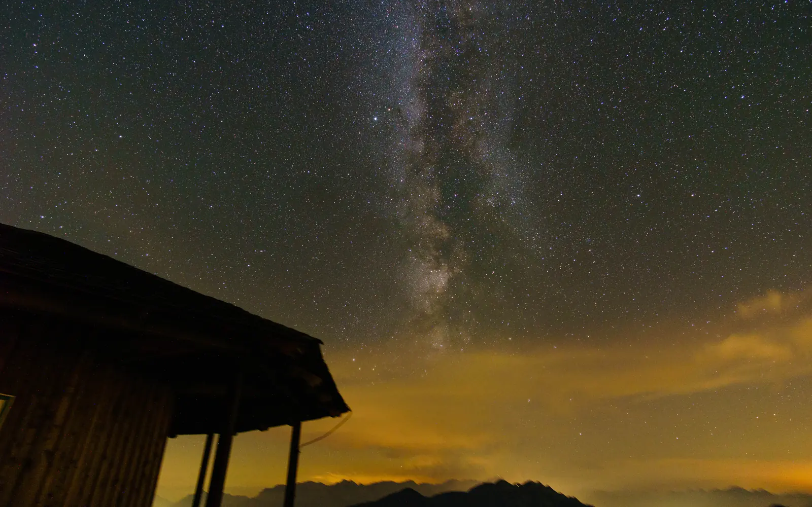 The Milky Way above a mountain hut, with glowing clouds in the valley below
